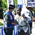 Two men talk on a blue pedestrian bridge at a cultural event one wears a blue uniform vest the other in traditional clothing carrying a large bag over his shoulder with a photo board and onlookers in the background