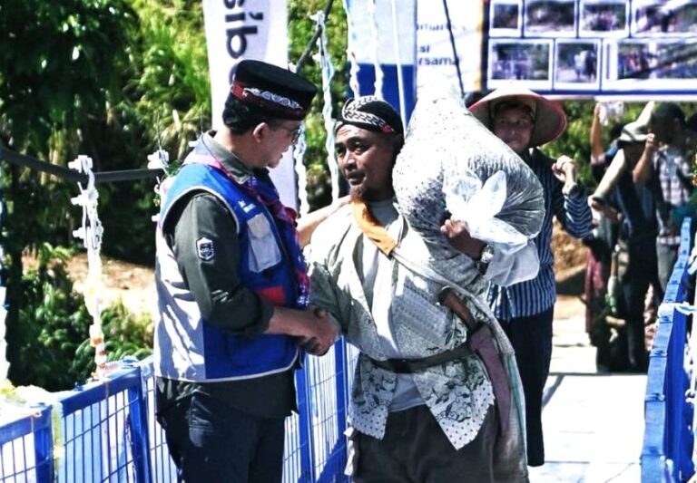 Two men talk on a blue pedestrian bridge at a cultural event; one wears a blue uniform vest, the other in traditional clothing carrying a large bag over his shoulder with a photo board and onlookers in the background.