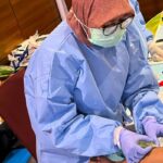 Healthcare worker in full ppe blue gown mask gloves drawing blood from a patients arm on a cluttered clinical table