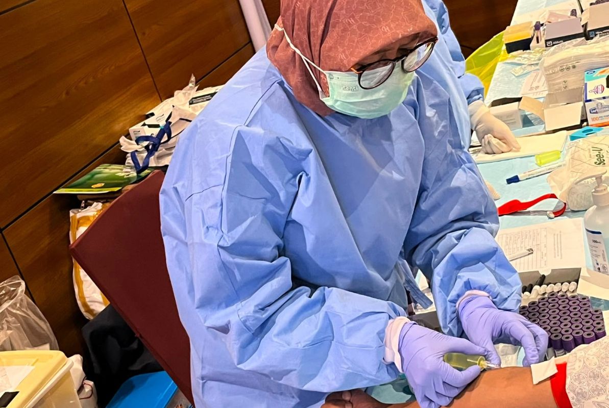 Healthcare worker in full ppe blue gown mask gloves drawing blood from a patients arm on a cluttered clinical table