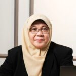 Professional woman wearing a beige hijab and glasses seated at a desk with a laptop in front of her