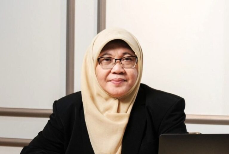 Professional woman wearing a beige hijab and glasses, seated at a desk with a laptop in front of her.