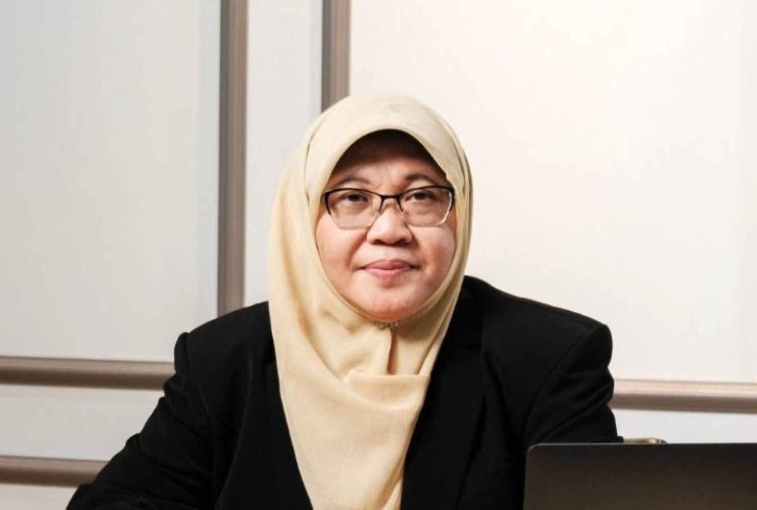 Professional woman wearing a beige hijab and glasses seated at a desk with a laptop in front of her