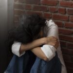 Person sitting on the floor with head bowed and arms wrapped around their knees beside a brick wall conveying sadness or distress