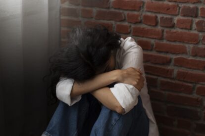 Person sitting on the floor with head bowed and arms wrapped around their knees beside a brick wall, conveying sadness or distress.