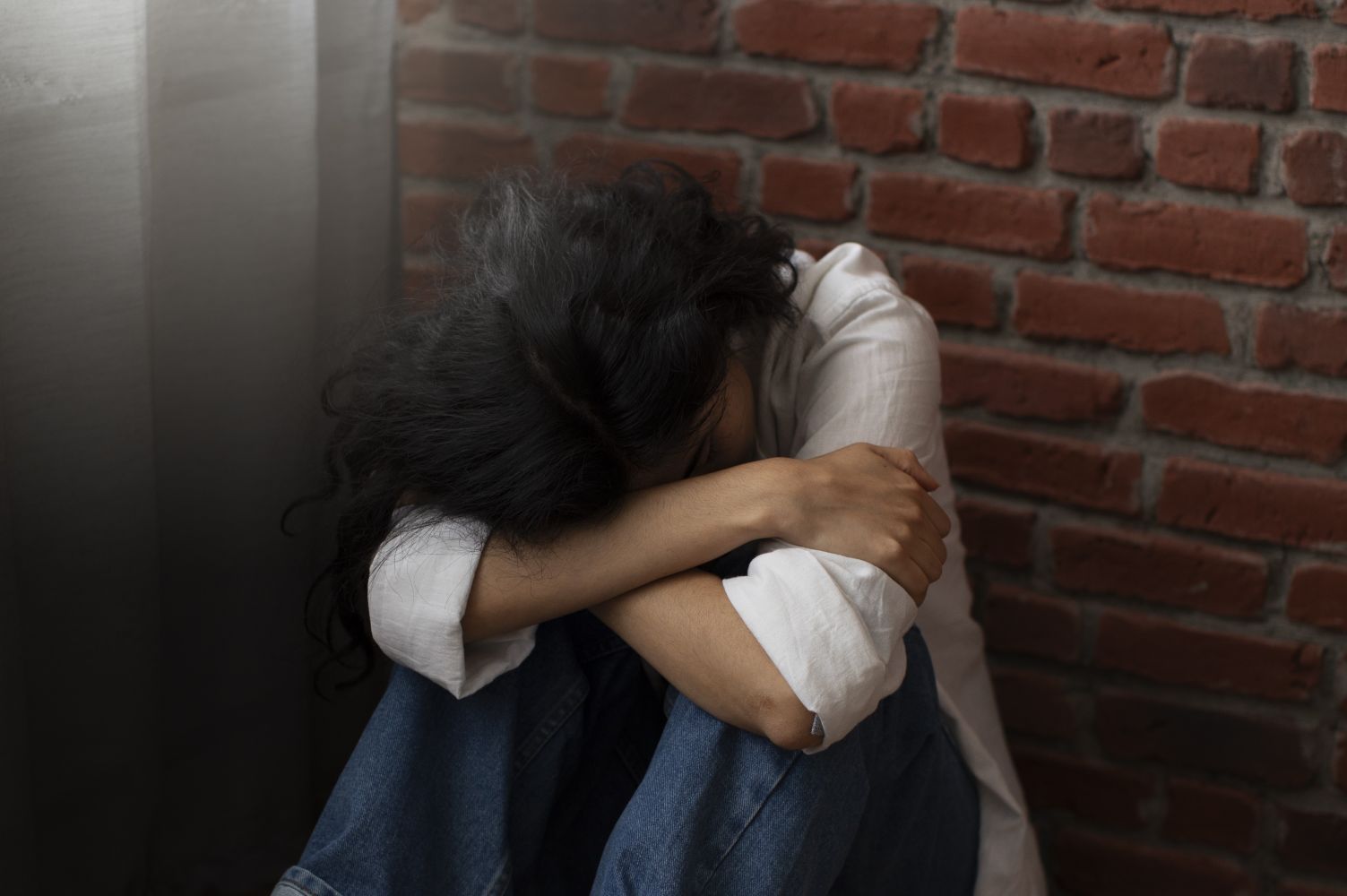 3gkorban catrawarta Person sitting on the floor with head bowed and arms wrapped around their knees beside a brick wall conveying sadness or distress