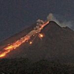 Volcano erupting at night with lava flowing down its slope and a small plume of smoke