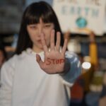 Person raises a hand toward the camera with stop written in red on the palm while a blurred crowd is in the background protest context