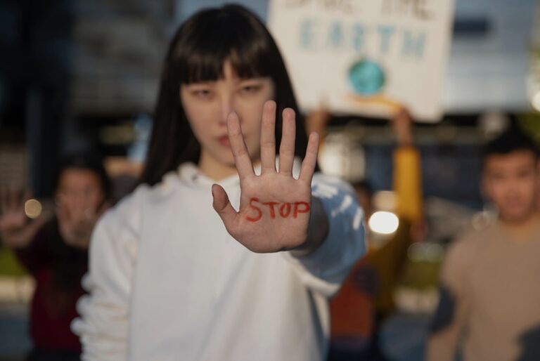 Person raises a hand toward the camera with 'STOP' written in red on the palm, while a blurred crowd is in the background (protest context).