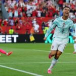 Soccer player in portugals white and green kit celebrates a goal on the field with a pink clad goalkeeper on the ground behind him and a cheering crowd in the stands behind
