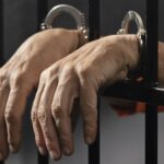 Hands in metal handcuffs gripping a jail cell bars orange jumpsuit visible in background