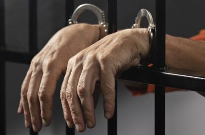 Hands in metal handcuffs gripping a jail cell bars, orange jumpsuit visible in background.