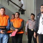 Two men in orange jail vests numbers 61 and 167 stand with security officers inside a building one holding a pink folder other people nearby
