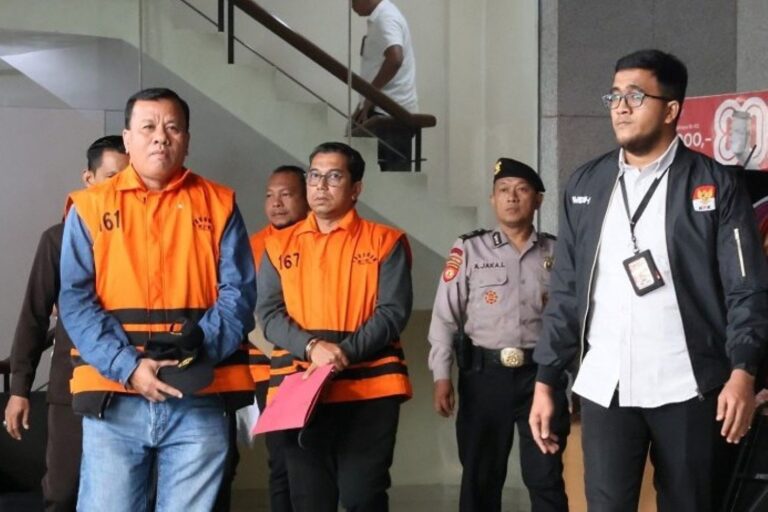 Two men in orange jail vests (numbers 61 and 167) stand with security officers inside a building, one holding a pink folder; other people nearby.