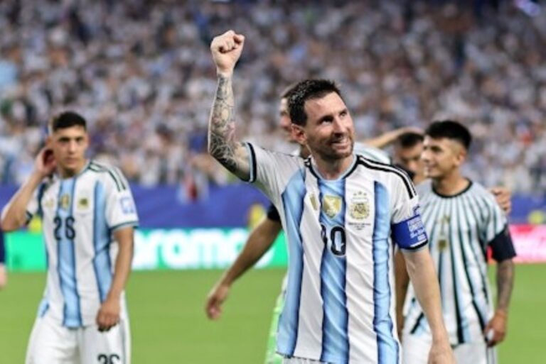 Argentine soccer players celebrate on the field as the captain raises a clenched fist in triumph, crowd in the background