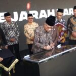 Official signs a document on a glass topped podium during a formal ceremony with several men in batik and traditional caps standing behind him