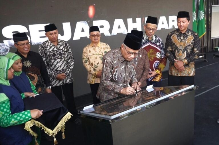 Official signs a document on a glass-topped podium during a formal ceremony with several men in batik and traditional caps standing behind him.
