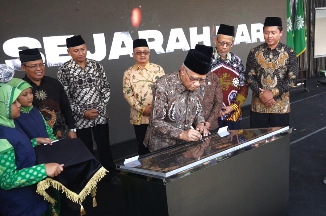 Official signs a document on a glass topped podium during a formal ceremony with several men in batik and traditional caps standing behind him