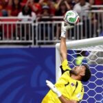 Soccer goalkeeper in a yellow kit reaches up to punch away a white green soccer ball near the goal