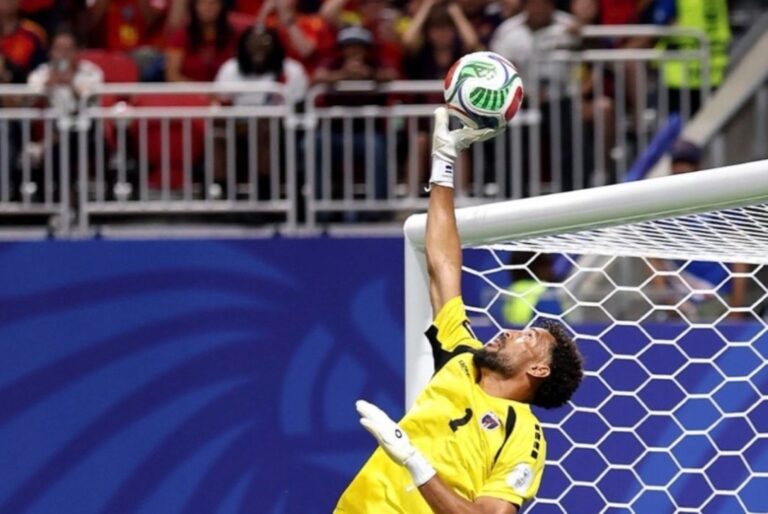 Soccer goalkeeper in a yellow kit reaches up to punch away a white-green soccer ball near the goal.