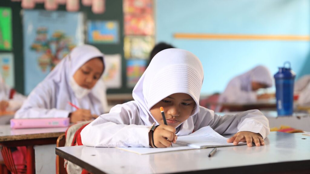 Young girl in a white hijab writes at a desk in a classroom focused on her homework with a pencil in hand and a blue water bottle in the background
