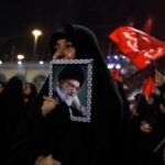 A woman in a black hijab holds a framed portrait of a bearded elderly man wearing a turban at a nighttime crowd rally with red flags in the background
