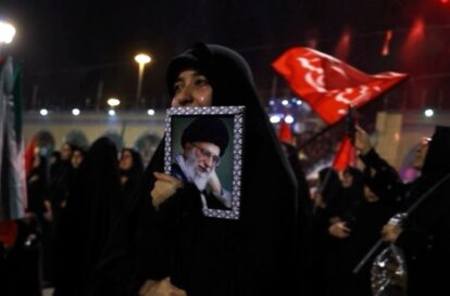 A woman in a black hijab holds a framed portrait of a bearded elderly man wearing a turban at a nighttime crowd rally with red flags in the background