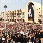 Huge crowd gathered in front of a beige arched building with a giant portrait of a cleric waving many people with flags nearby