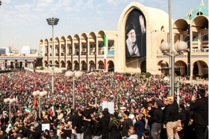 Huge crowd gathered in front of a beige arched building with a giant portrait of a cleric waving, many people with flags nearby.