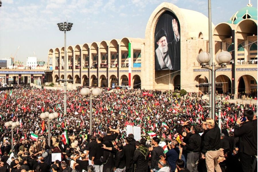 5gmakam1 catrawarta Huge crowd gathered in front of a beige arched building with a giant portrait of a cleric waving many people with flags nearby