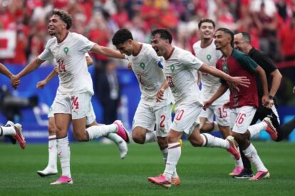 Morocco national team players celebrate a goal, running across the field in white kits with green badges.
