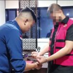 Two men are indoors a man in a blue shirt helps a man wearing a pink and black vest put on a bracelet at a desk