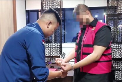 Two men are indoors; a man in a blue shirt helps a man wearing a pink and black vest put on a bracelet at a desk.