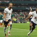 England soccer players celebrate a goal on the pitch wearing white jerseys with numbers 9 and 10 with fans in the stands behind them