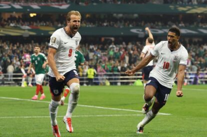 England soccer players celebrate a goal on the pitch, wearing white jerseys with numbers 9 and 10, with fans in the stands behind them.