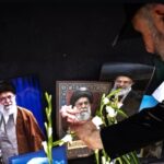 Man in a black shirt places white flowers in front of framed portraits of iranian clerics against a dark background as part of a memorial display