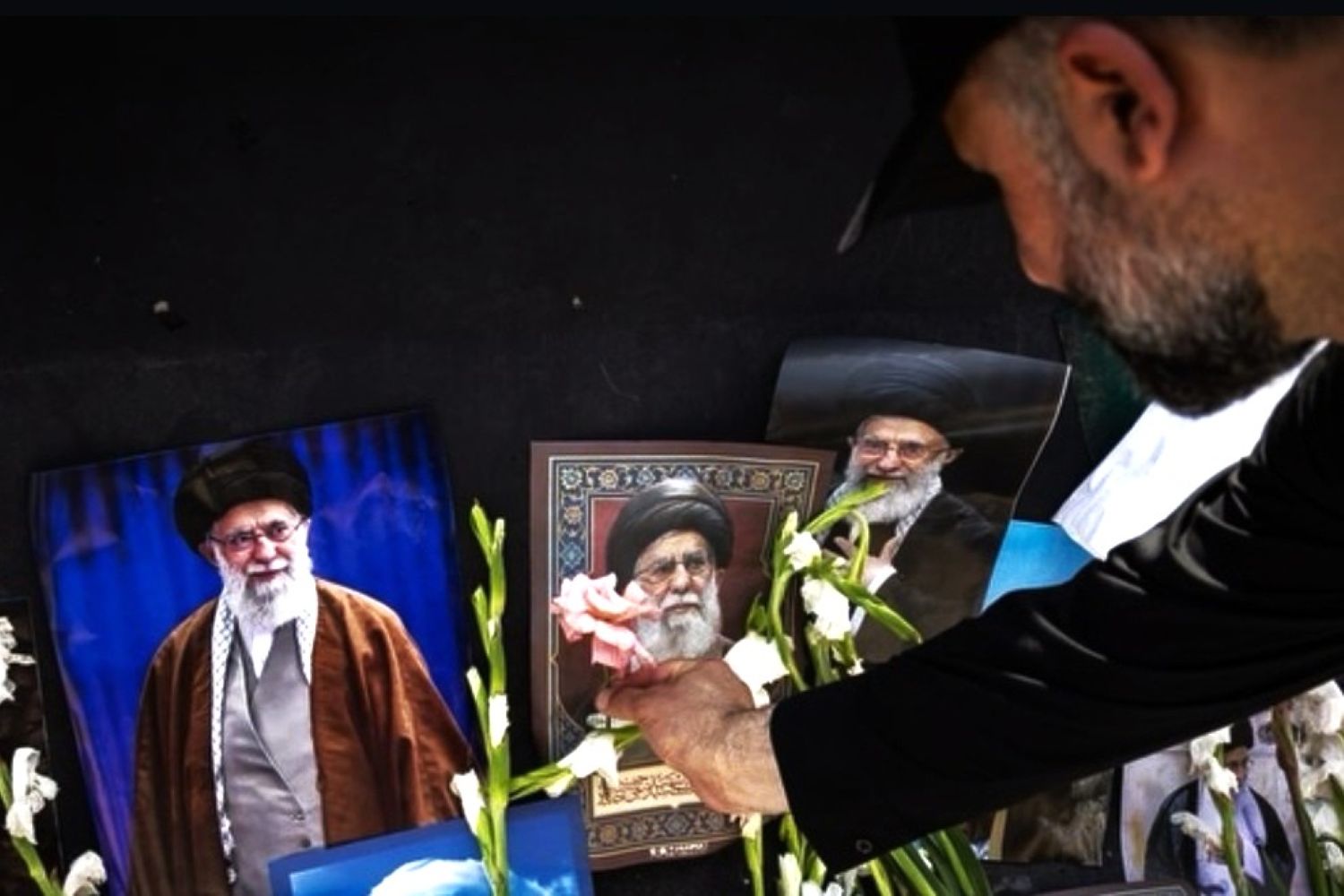 6giran catrawarta Man in a black shirt places white flowers in front of framed portraits of iranian clerics against a dark background as part of a memorial display