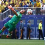 Soccer goalkeeper in a green kit dives to save a ball near the goal as a crowd watches