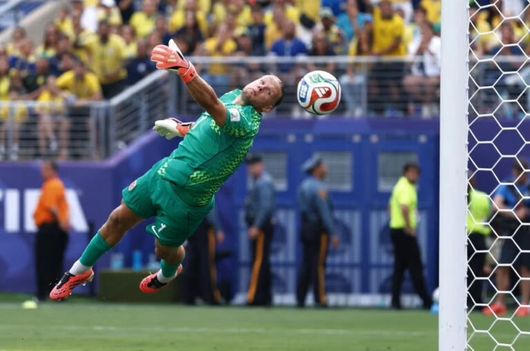 Soccer goalkeeper in a green kit dives to save a ball near the goal as a crowd watches.