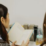 Two people sit side by side reading an open book at a desk with metal bookends and a mason jar drink nearby