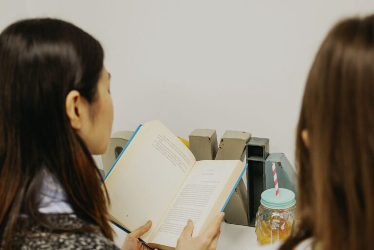 Two people sit side by side reading an open book at a desk, with metal bookends and a mason jar drink nearby.
