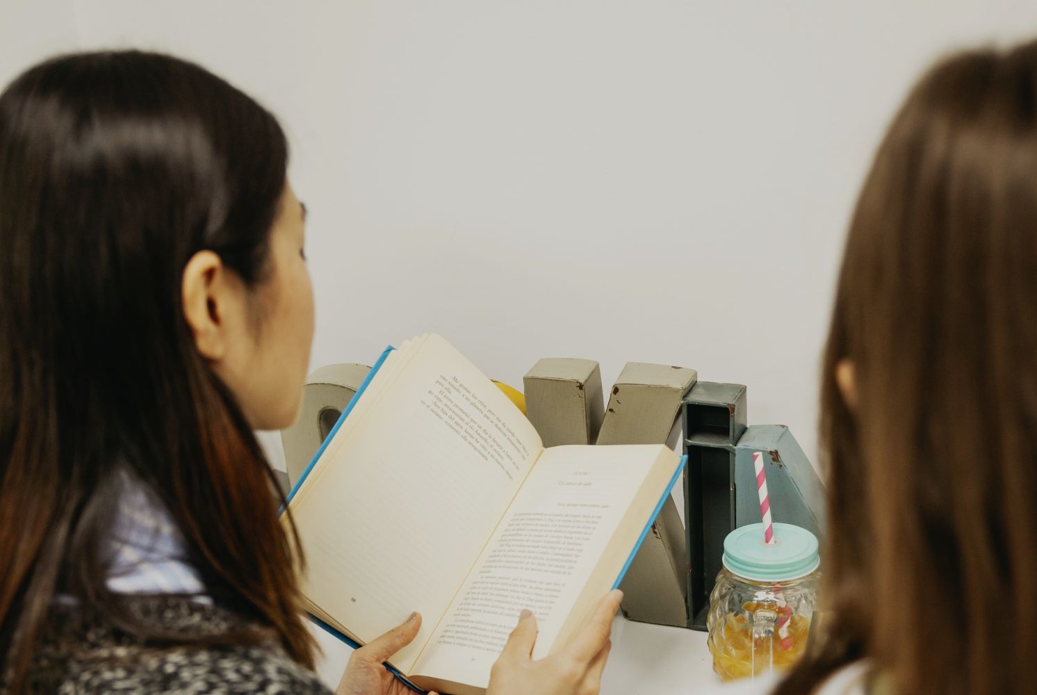 6gliterasi catrawarta Two people sit side by side reading an open book at a desk with metal bookends and a mason jar drink nearby
