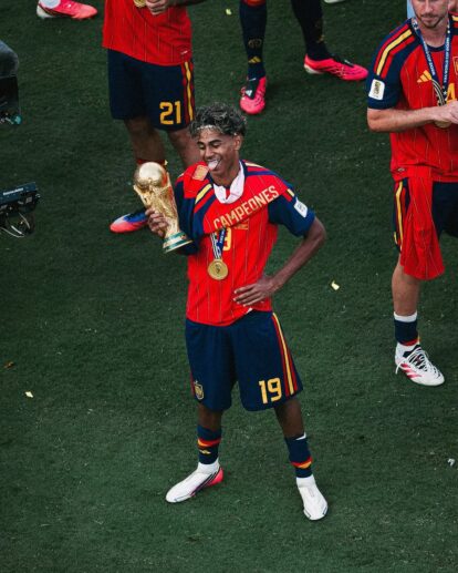 Soccer player in red jersey with 'CAMPEONES' holding a golden trophy and wearing a gold medal, celebrating on the field.