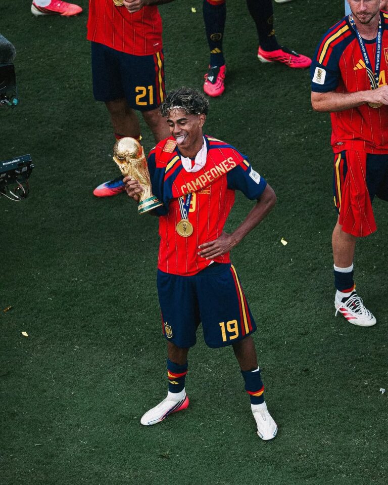 Soccer player in red jersey with 'CAMPEONES' holding a golden trophy and wearing a gold medal, celebrating on the field.