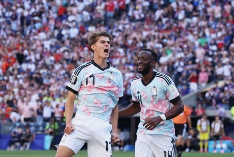 Two soccer players in pale pink camouflage jerseys celebrate a goal on the field as the crowd cheers.