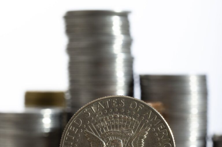 Close-up of a large silver coin in the foreground, with an eagle design, and blurred stacks of coins in the background.
