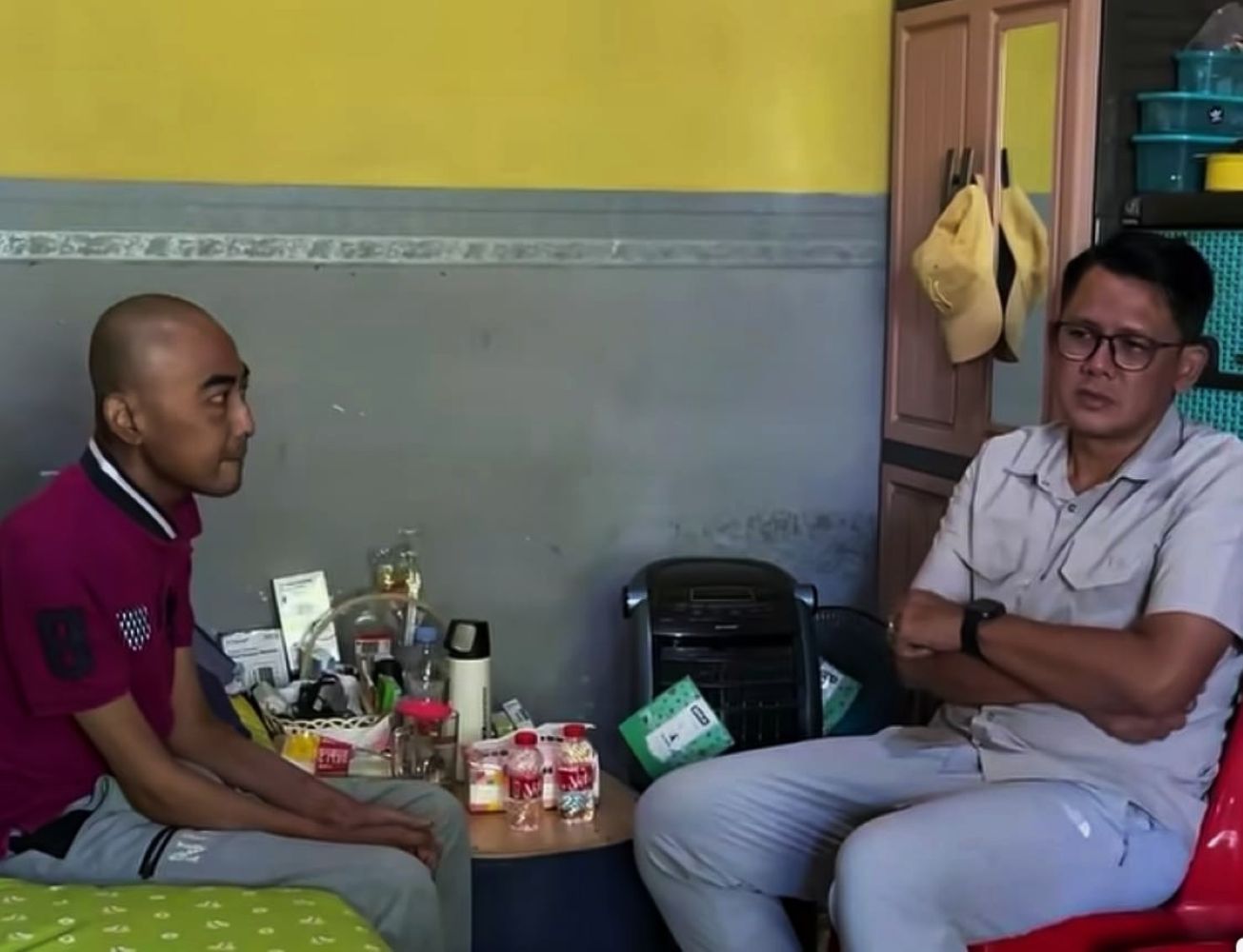 Two men sit facing each other in a small room a cluttered table with bottles and items sits between them A yellow wall and a closed closet with hats are in the background