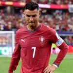 Portuguese footballer wearing red jersey with number 7 on a football pitch crowd in the stands behind him