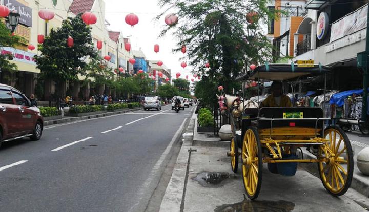 Kawasan jalan malioboro catrawarta Street in a town with red lanterns overhead storefronts on both sides and a yellow horse drawn carriage parked on the sidewalk