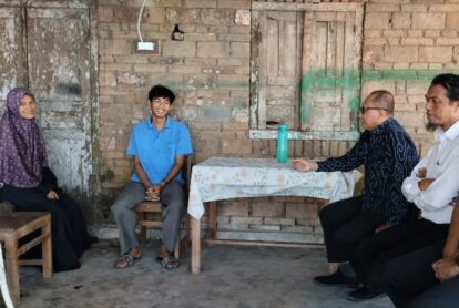 A young man in a blue polo sits smiling between a woman in a purple hijab and three men at a wooden table in a brick-walled room with a turquoise water bottle on the table.